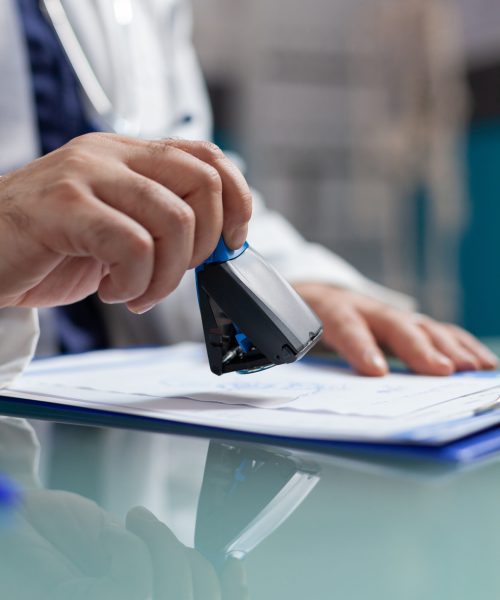 Doctor holding medical stamp to put on prescription paper at health care exam. Hand of physician putting seal on checkup report, giving medicine to patient at consultation. Close up